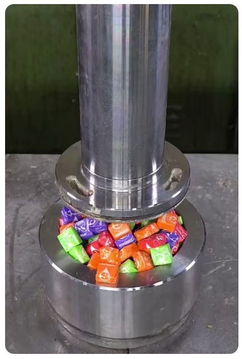Wrapped colorful candies packed in a metal cup under a hydraulic press
